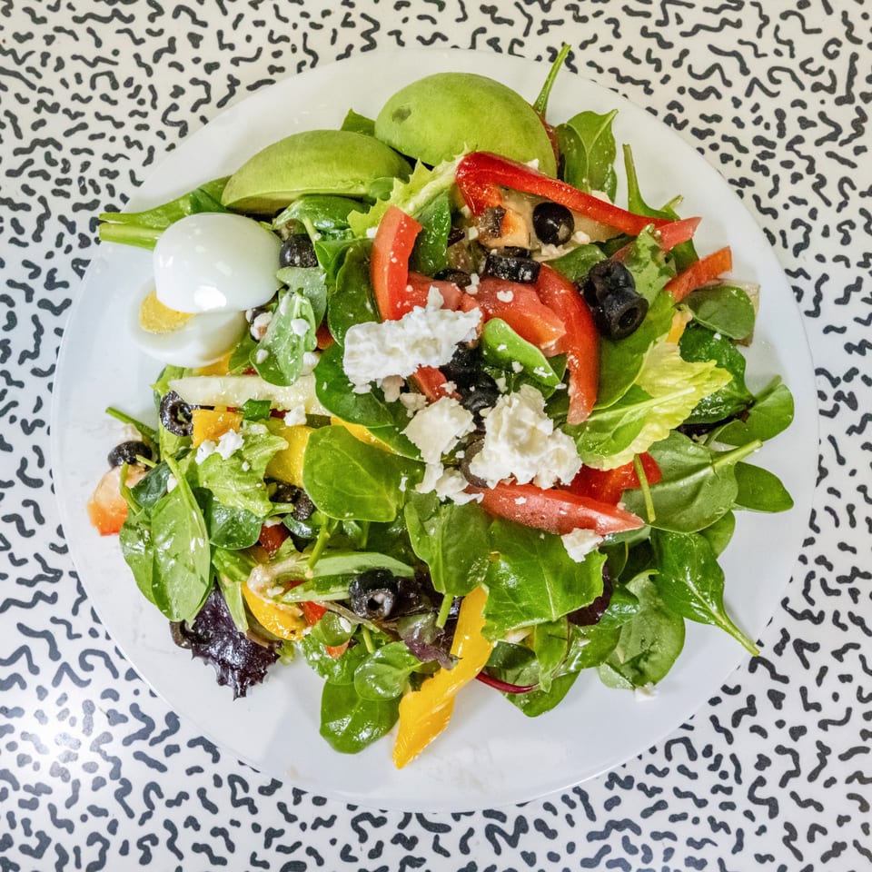 Fresh salad with spinach, tomatoes, olives, feta, avocado, and a hard-boiled egg on a white plate.